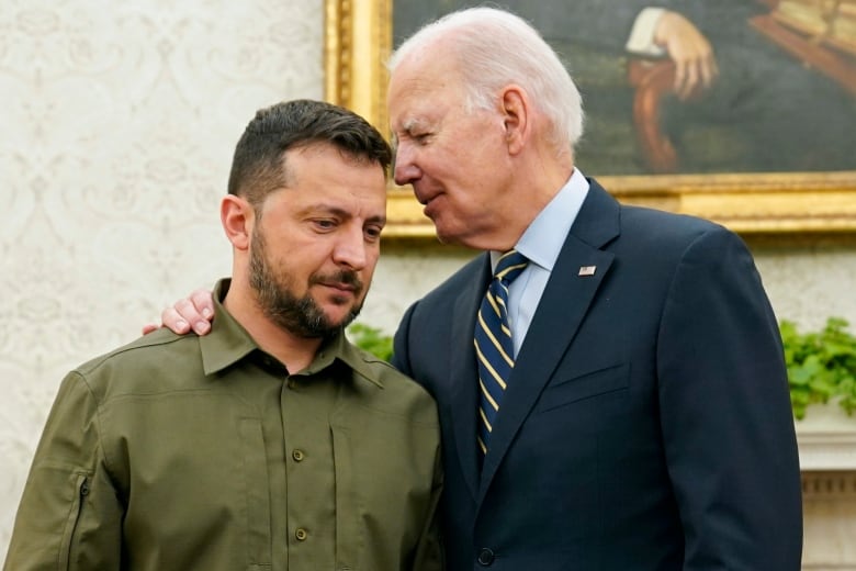 A taller grey-haired man in a suit, puts his hand on the shoulder of a shorter, bearded man in a green button-up shirt. 