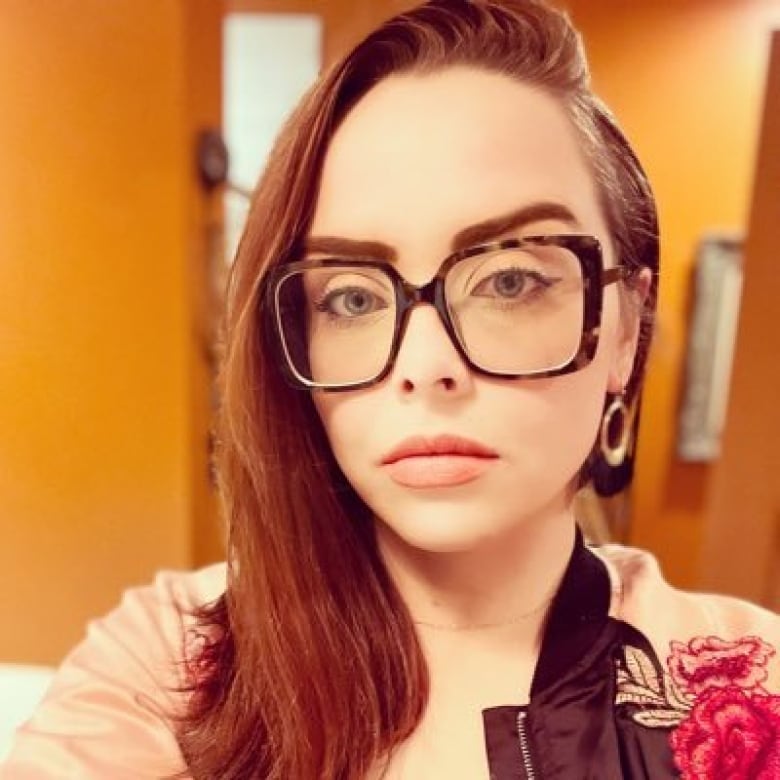 The nurse Amy Hamm is shown in an orange-toned selfie. She is white with long brown hair and large, dark-rimmed glasses.