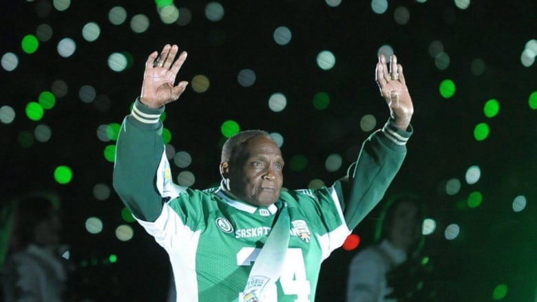 A man wearing a football jersey waves to fans in the stands with both arms in the air.