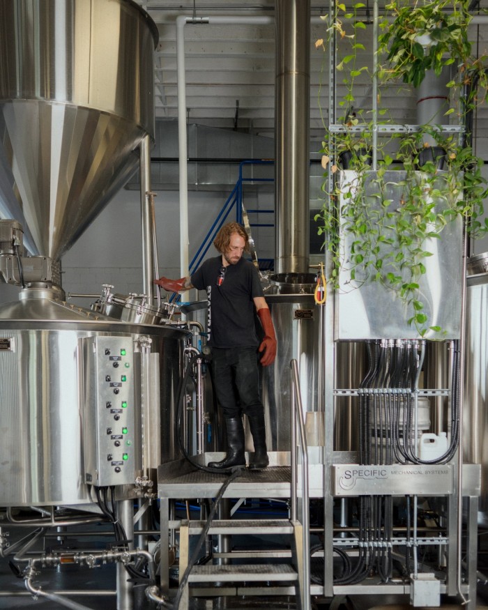 Brewer Ben Owens in a boiler suit and long red rubber gloves standing between two steel vats at Superflux brewery