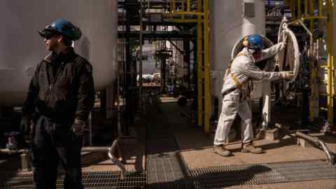 Two men in coveralls and hard hats stand beside industrial storage tanks