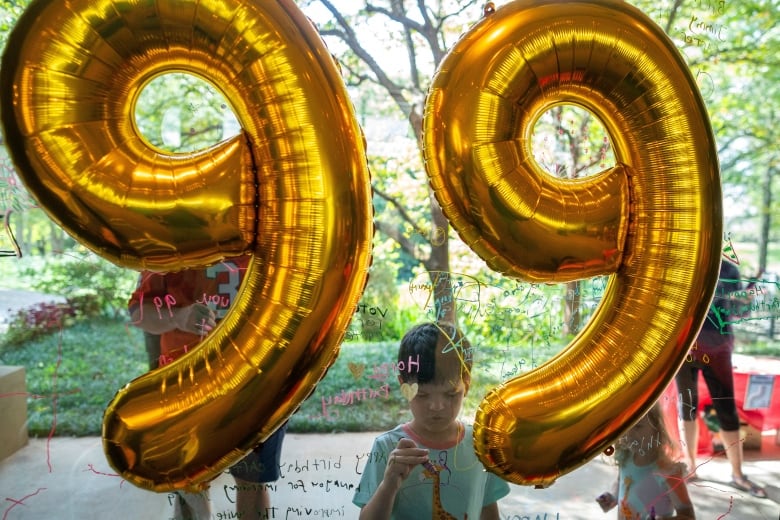 A child writes a message on a window as balloons shaped as the number 99 is seen in the foreground.