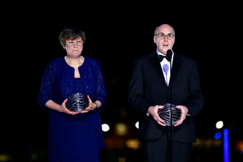 A man and a woman in formal wear are shown on a stage with a dark background, each holding an award in their hands.