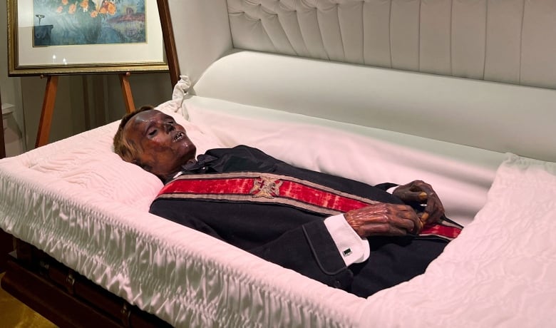 A browned, mummified corpse wearing a suit and a red sash lying in a casket. 