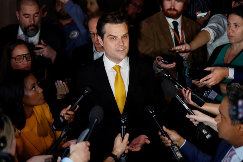 Dark haired man in dark suit and yellow tie speaks surrounded by reporters