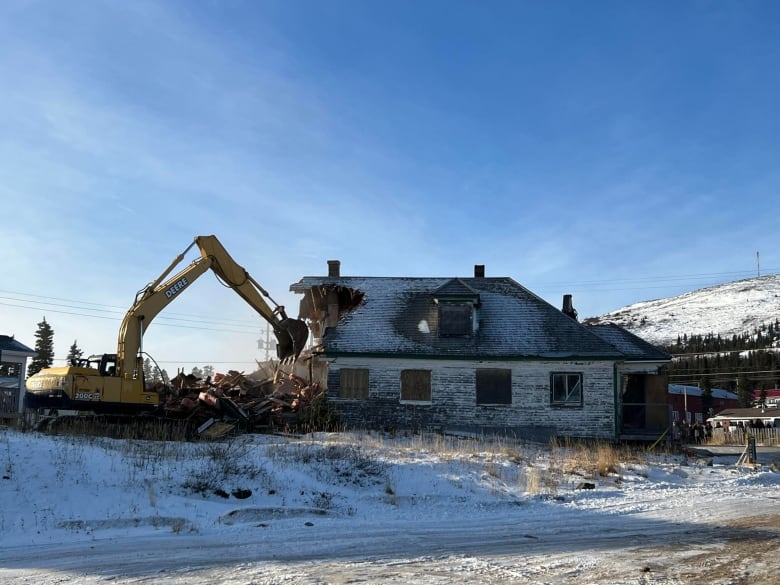 An excavator cuts through an abandoned building. It is about a third of the way through demolishing it. 