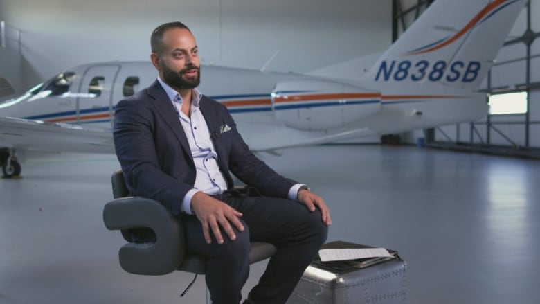 A person sits on a chair in an airplane hanger in front of a small plane.