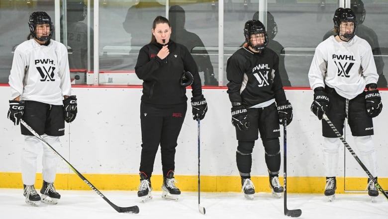 A female hockey coach blows a whistle on the ice, where she stands between three players. Two players are wearing white jerseys and one player is wearing a black jersey, all with PWHL written on them.