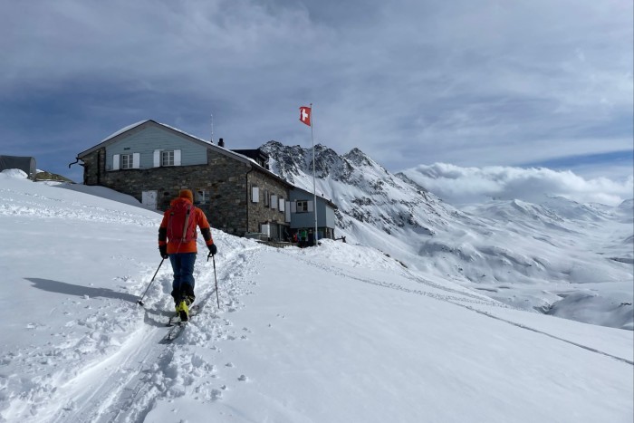 A skier in red heads uphill through thick snow towards a chalet or mountain lodge where a Swiss flag is flying