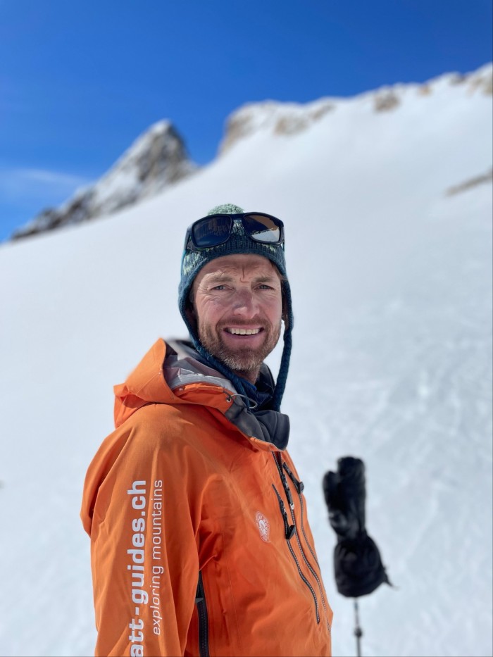 A rugged-looking male ski guide wearing an orange jacket and ski goggles grins at the camera