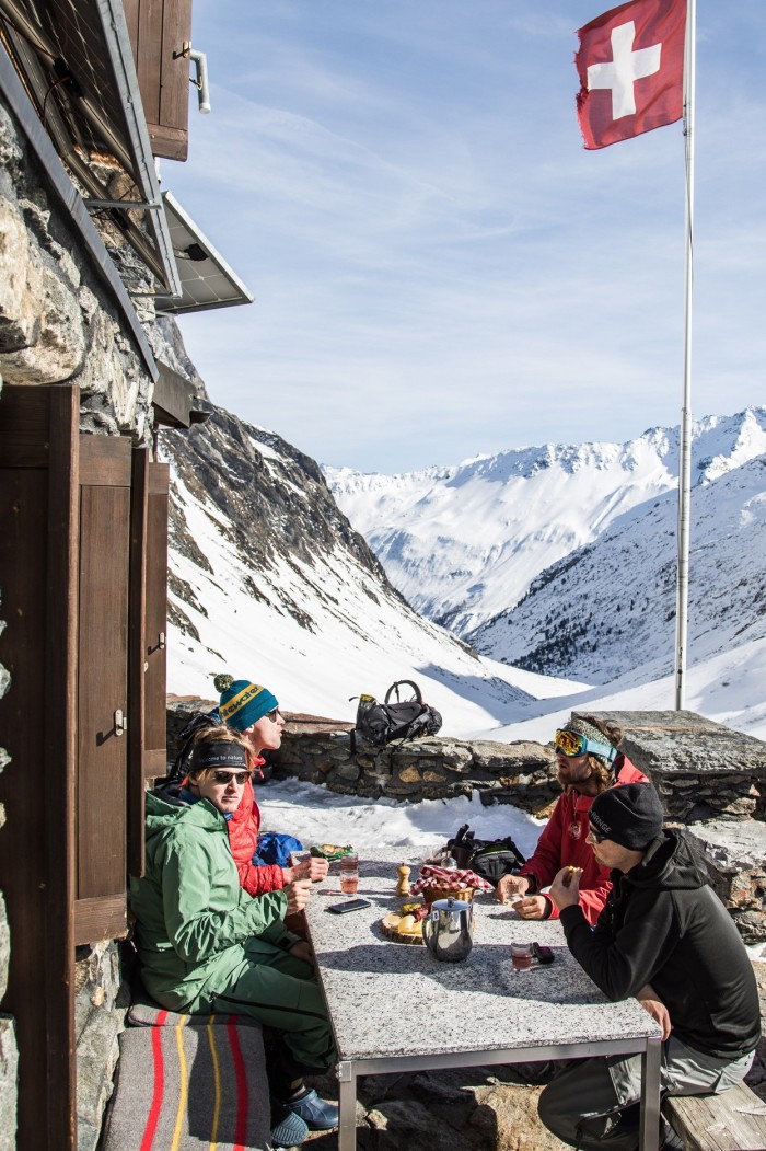 A group of skiers seated at a table outside with a snowy backdrop and a Swiss flag flying