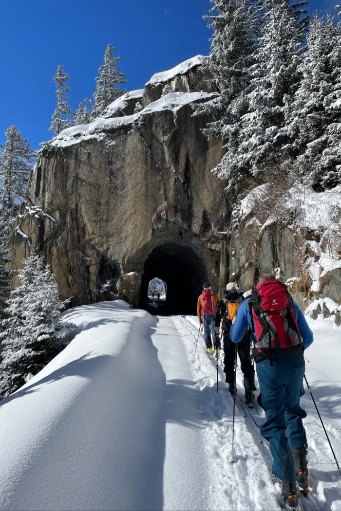 Three skiers cross-country ski into a small tunnel carved out of a mountainside