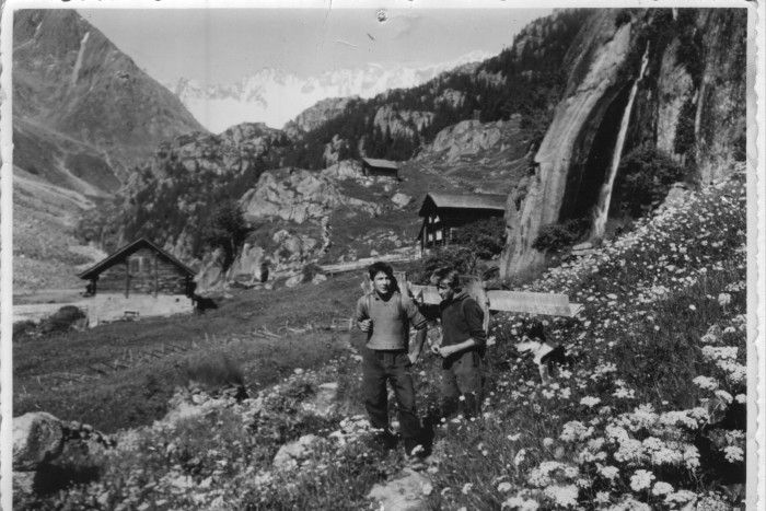 A black-and-white photograph circa 1940s of two boys hiking on an Alpine mountainside in spring, with flowers in the foreground and snowy peaks in the background