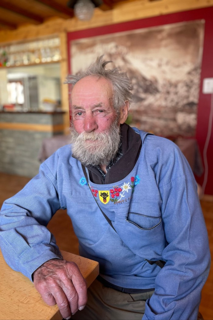 An elderly man with a thick long beard wearing a blue smock embroidered with flowers leans on a table, looking at the camera