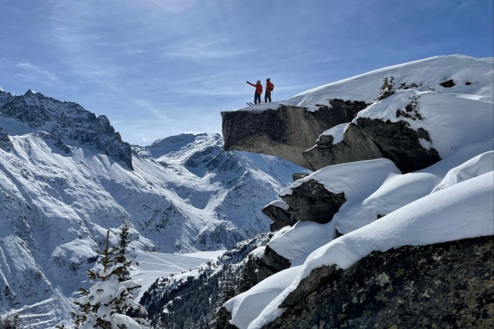 Two skiers in red stand on a precarious-looking outcrop of rock in a snowy mountainscape with blue skies behind them