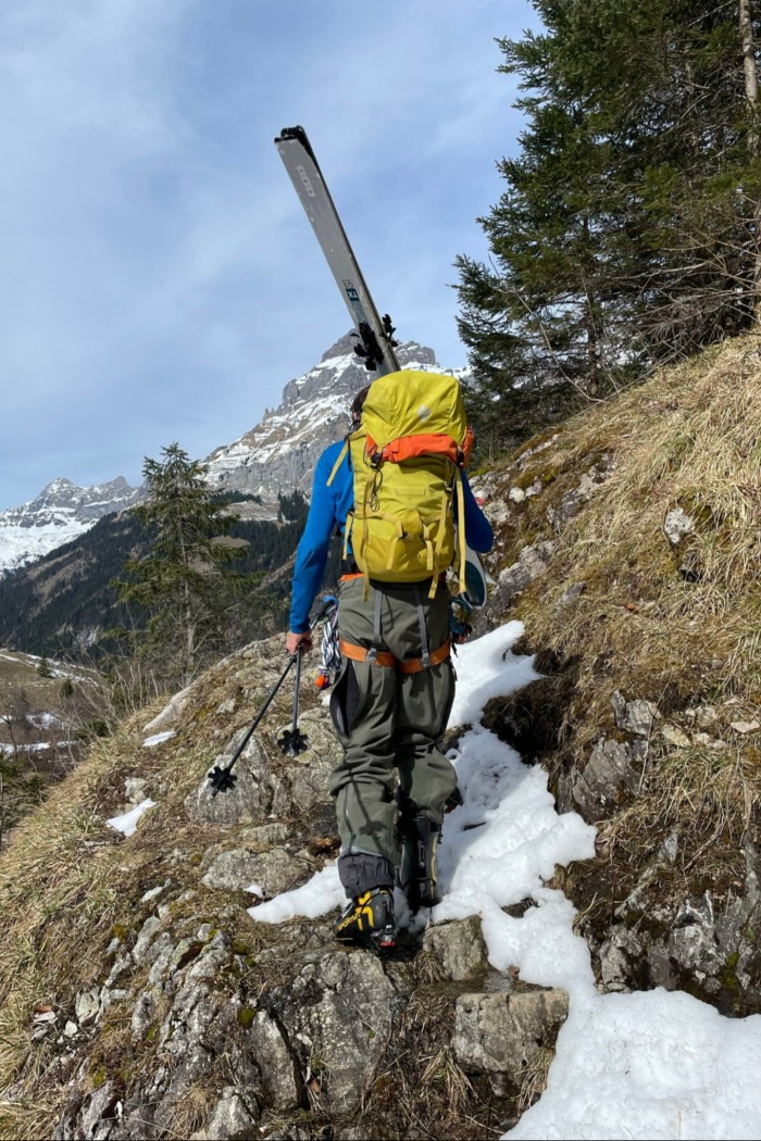 A man carrying skis stands, back to the camera, on a rocky slope with only patches of snow