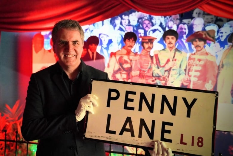 A smiling gray-haired man in a suit wears white gloves and holds up a Penny Lane street sign. Behind him is a picture of The Beatles.