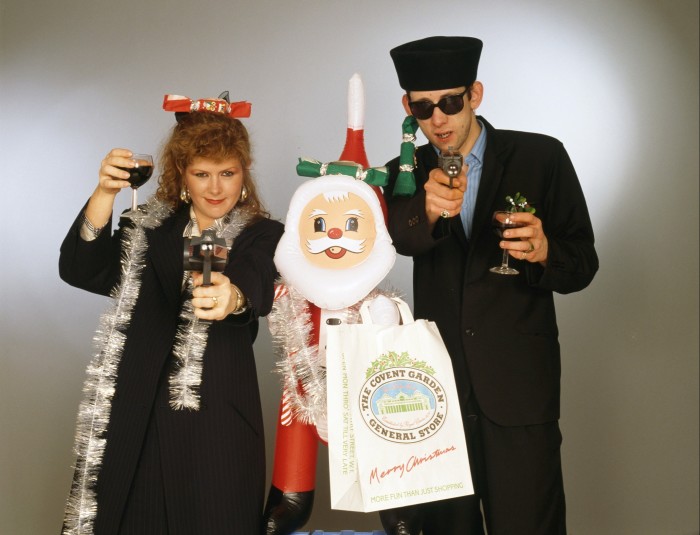 A woman wearing silver tinsel and holding a glass of wine and a man holding a glass of wine stand either side of an inflatable Santa Claus