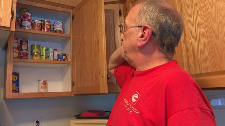 A man wearing a red t-shirt looks inside an open kitchen cupboard filled with canned food.