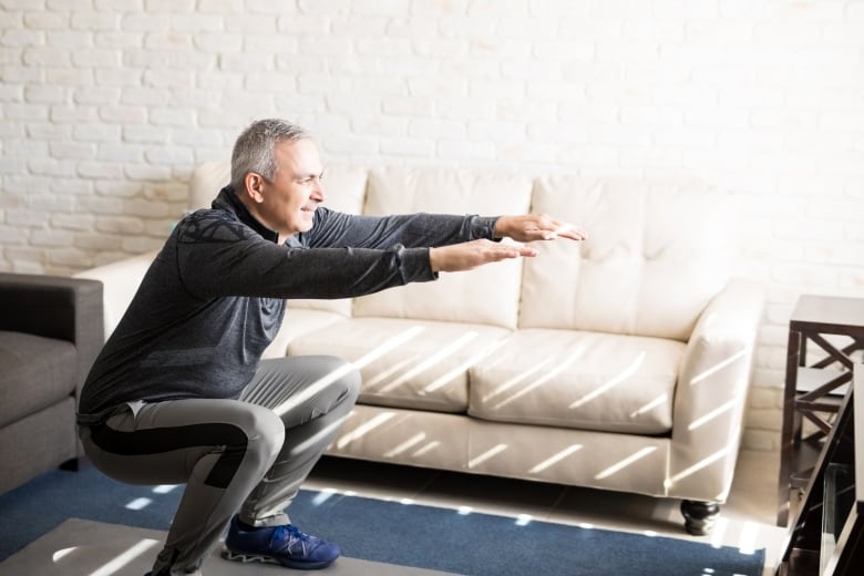 A man does squats wearing exercise clothes on an exercise mat in his living room. 