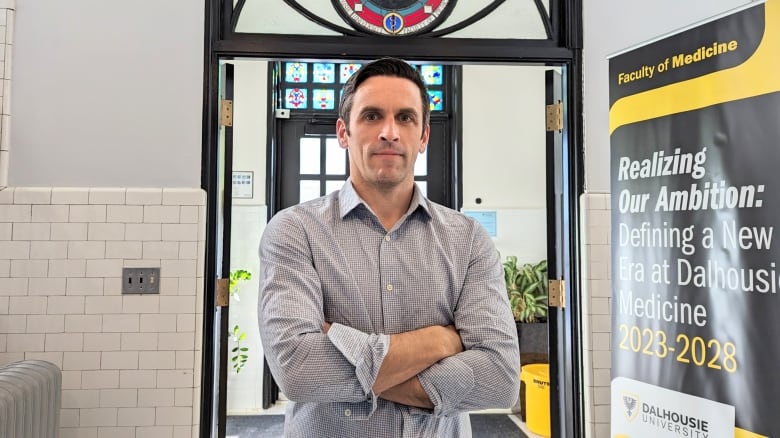 A man with short hair, crossed arms and a neutral expression. He is wearing a collared shirt and stands in front of a door with stained glass.