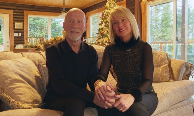 A woman and a man hold hands on their couch, in front of a Christmas tree.