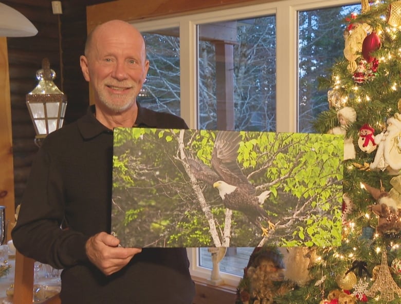 A man smiles and holds a large mounted photo of an eagle. He stands in front of his Christmas tree.