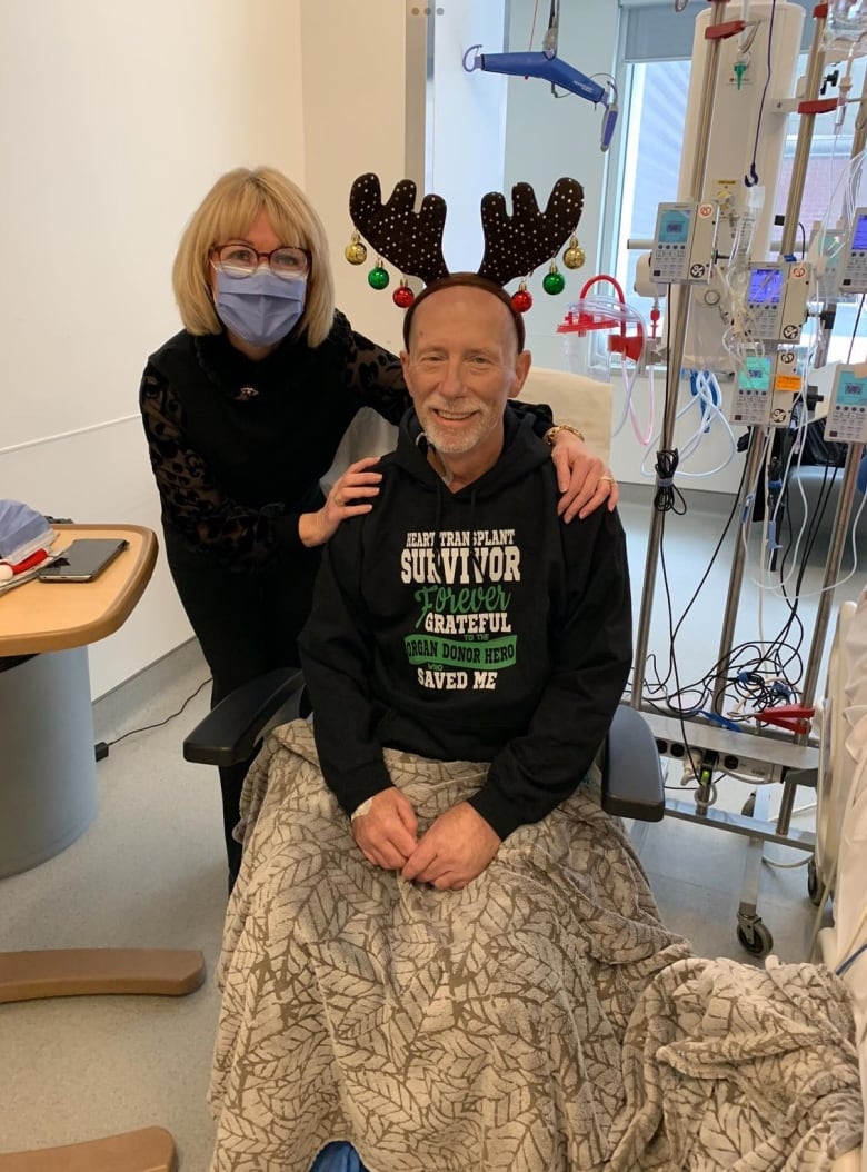 In a hospital, a woman in a mask stands with her hands on the shoulder of a man in a chair, who is wearing a reindeer antler headband.  