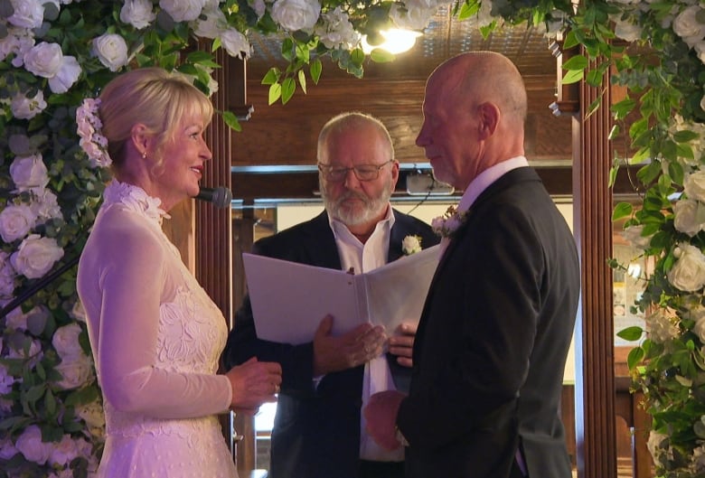 A bride in a white dress and a groom in a black suit stand in front of an officiant, surrounded by flowers.