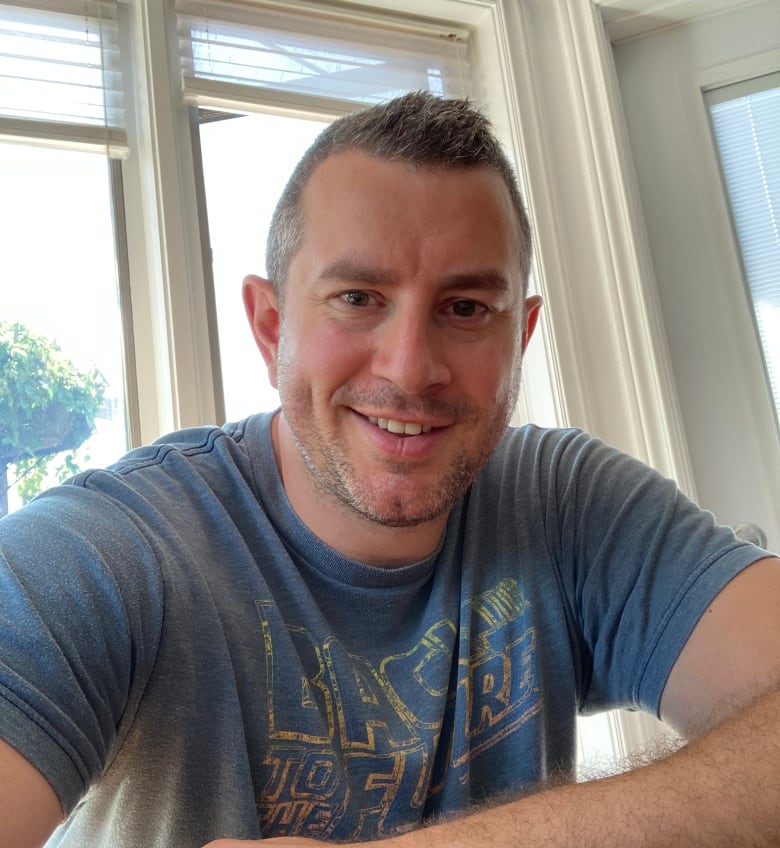 A man with short greying hair and a stubbly beard wears a faded blue "Back to the Future" t-shirt and sits looking into the camera. Behind him, there are large windows with white moulding.