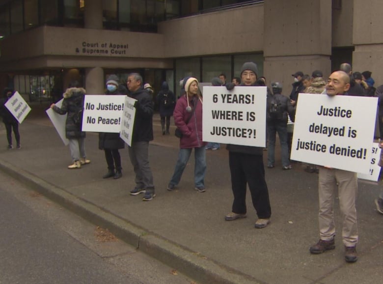 A group of people hold signs on a sidewalk with messages including "Justice delayed is justice denied!", "6 YEARS! WHERE IS THE JUSTICE?!" and "No Justice! No Peace!: