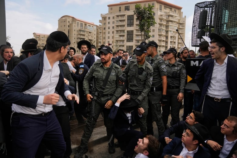 Ultra-Orthodox Jewish men wearing suits, skullcaps and fedoras sit and stand in the middle of a road and shout as some struggle with men and women wearing Israeli military uniforms. 