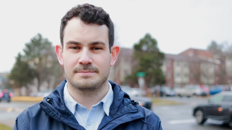 A man with a shaved mustache and beard has a neutral expression. He's wearing a collared shirt and a windbreaker. Behind him is a street and cars.