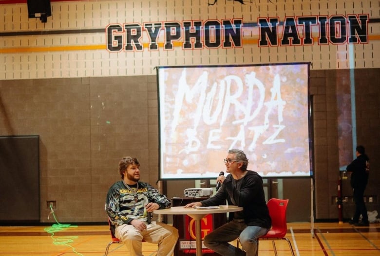 Two people sit at a raided table in a gymnasium. "Murda Beatz" is projected on a screen behind them. 