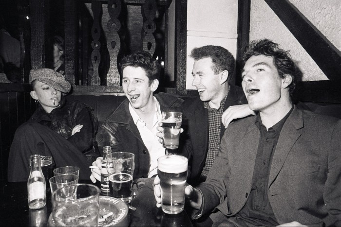 Black and white photo of three men lifting their pint glasses in a pub while a woman in a flat cap, smoking, looks on