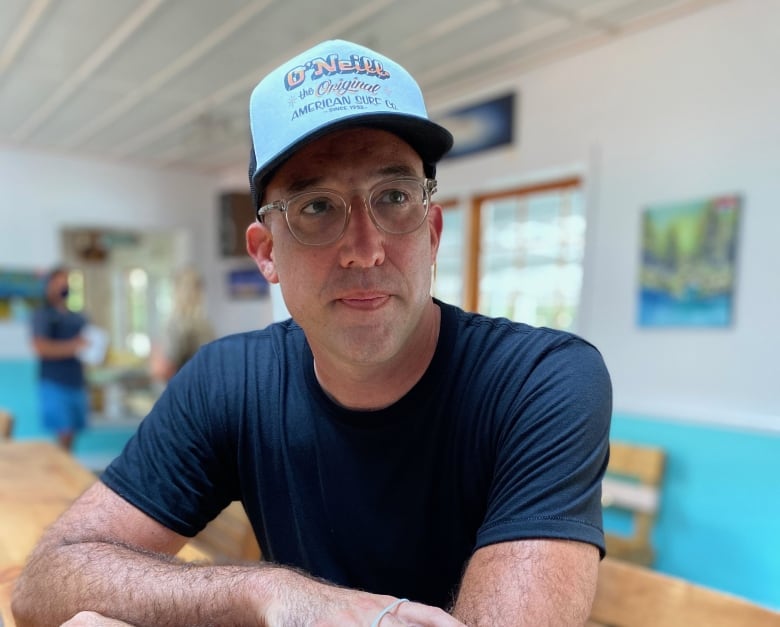 A man in a blue shirt, light blue hat and glasses sits at a table in a small restaurant.