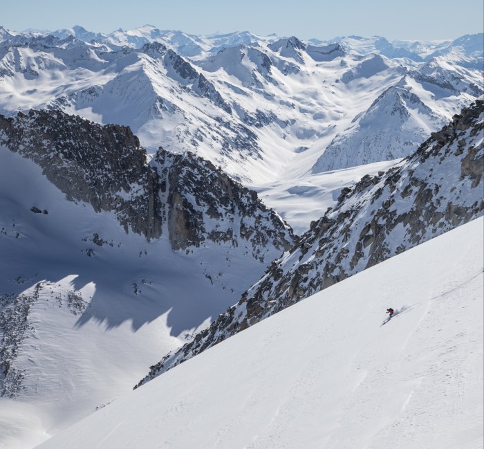 A lone skier in the foreground speeds down a steep slope, with craggy snowy mountains in the background