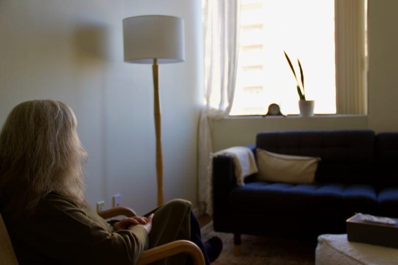 A person can be seen sitting in a chair facing a couch with the hands folded on their knee. 