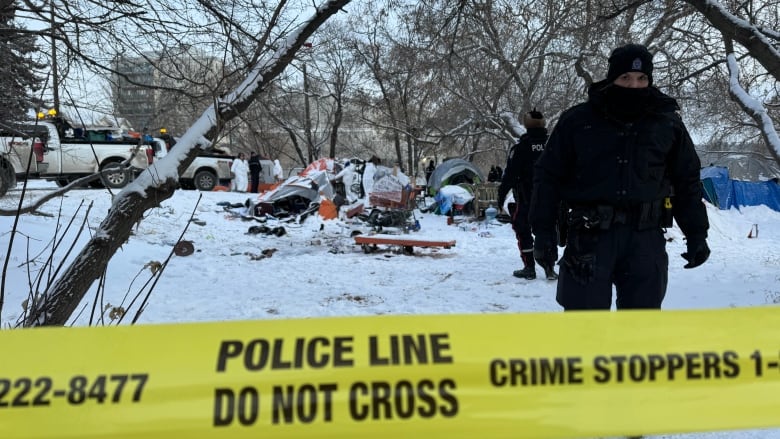 A police officer stands in the snow. There is a cluster of tents and tarps in the background