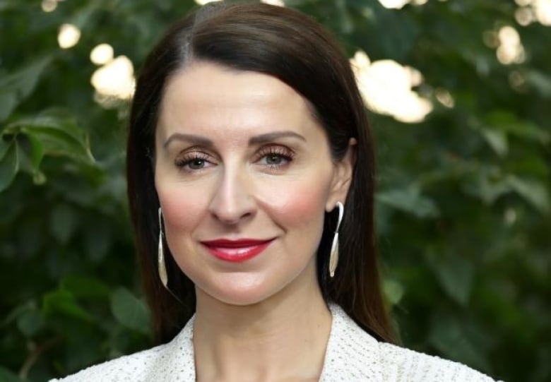 A woman with long hair, a blazer, and earrings smiles into the camera. 