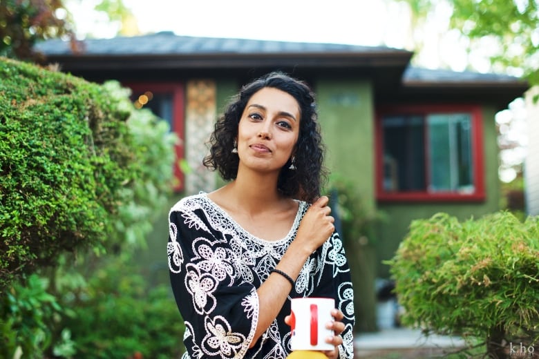 A woman holding a cup of tea stands in front of a house, framed by a green hedge. 