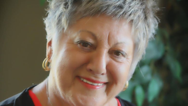 A close-up portrait of a smiling woman with short, greying hair, wearing red lipstick and gold earrings.