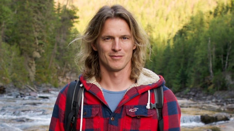 A man with shoulder-length blond hair and red, plaid jacket stands by a river and evergreen trees.