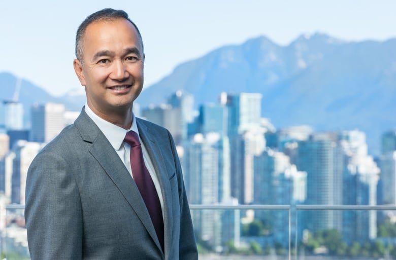 Profile photo of an adult Asian man wearing a grey suit in front of a city skyline of buildings with mountains in the background.