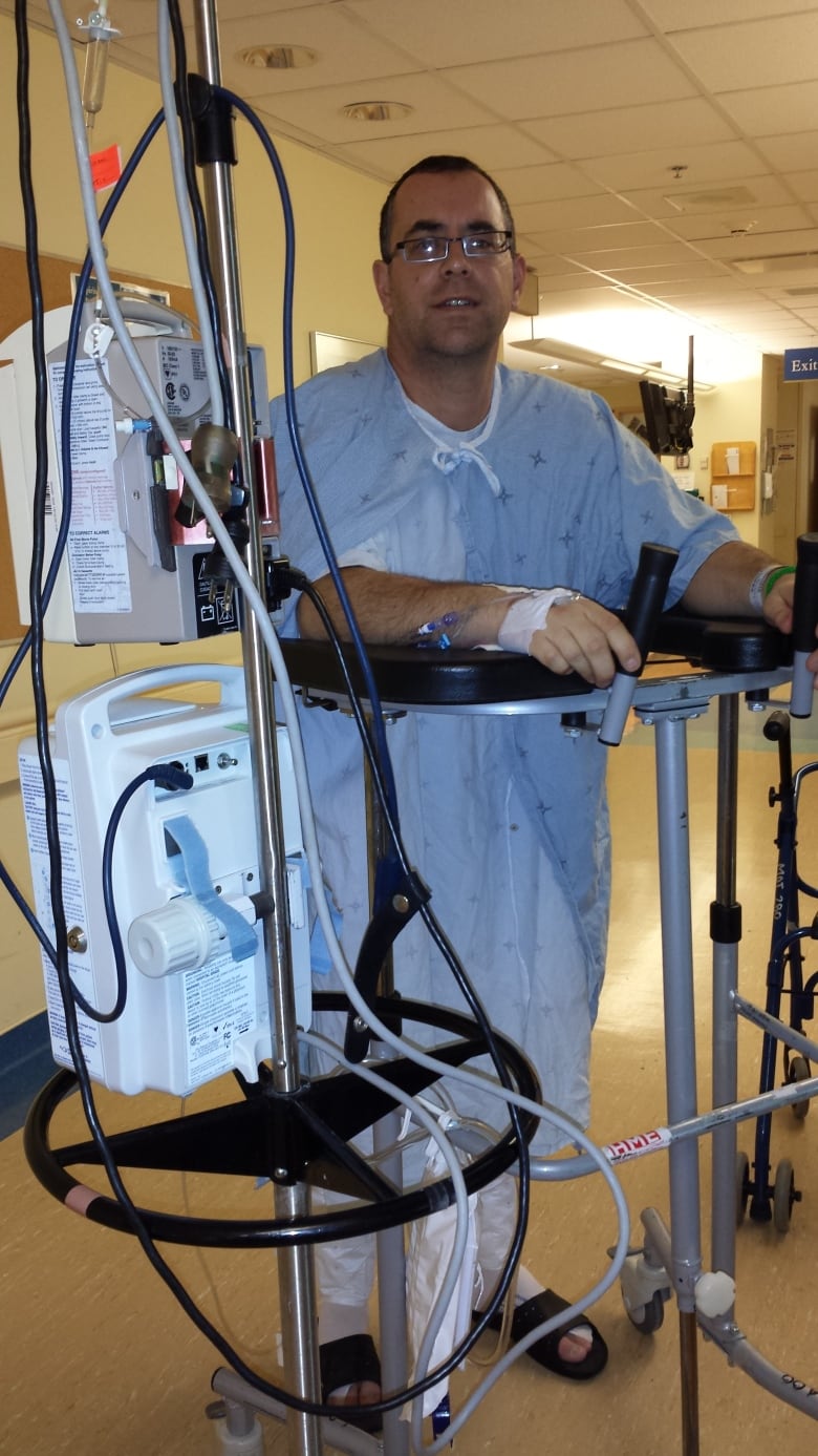 A man in a hospital gown stands, leaning on hospital equipment in a hospital hallway. He is white, middle-aged, wearing glasses.