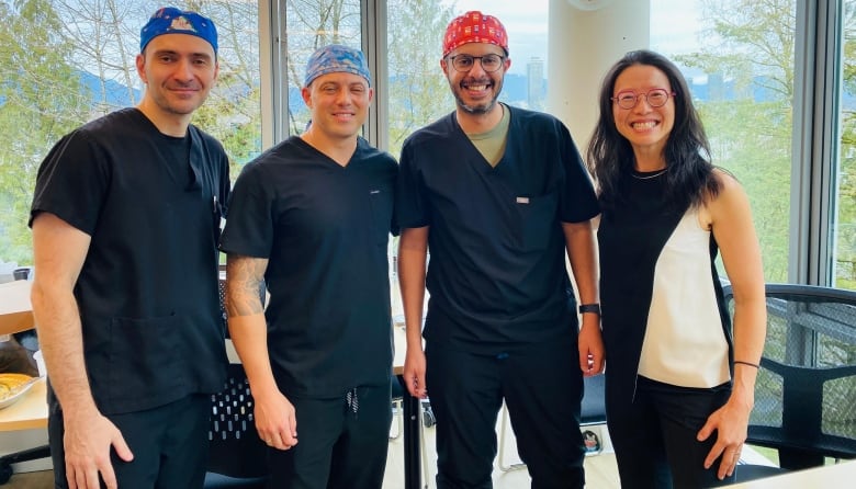 A group of four people — three men and one woman — stand together while smiling a the camera. The men are dressed in medical scrubs.