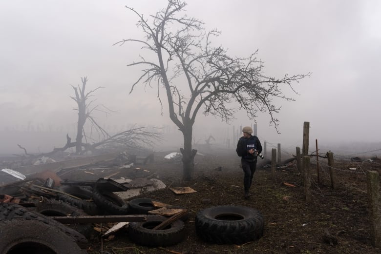 A man in a flack jacket, with the word "press" on it, walks through a misty dirty field littered with detritus. 