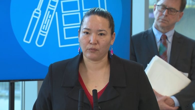 A First Nations woman, with her hair pulled back and warning colourful, dangling earrings speaks at a microphone at at Government of B.C. press conference.