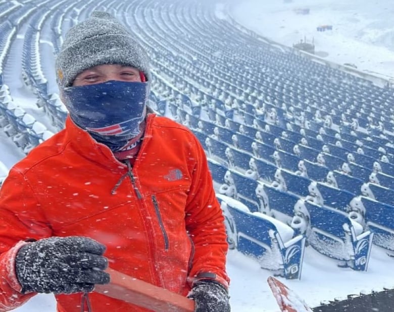 A man in a bright orange jacket and a New England Patriots face warmer holds a shovel while standing in an empty, snow-covered football stadium. 