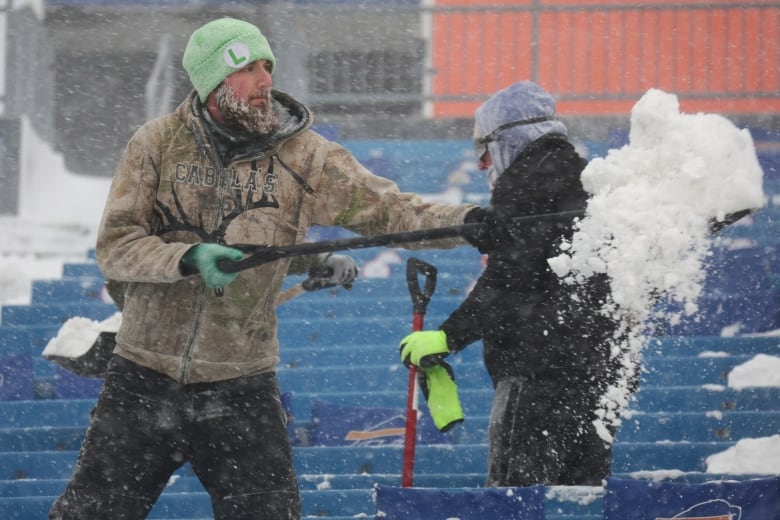 A man tosses snow in a shovel while standing amid the empty blue seats of a football stadium. Another person walks behind him, shovel in hand. 